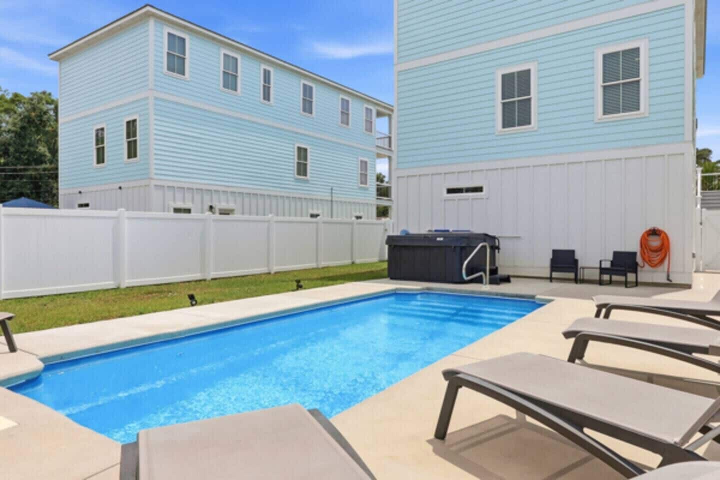 A swimming pool with lounge chairs and a hot tub in a backyard with a white fence and two-story houses.