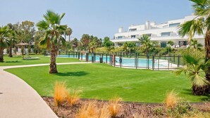 Zona de piscina con piscina cercada, palmeras y un edificio moderno al fondo.