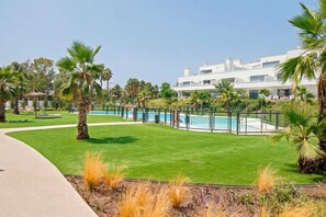 A pool area with a fenced pool, palm trees, and a modern building in the background. - Corner property-free parking-Pool-close to beach - Acgua Garden B-1-BA (Marbella)