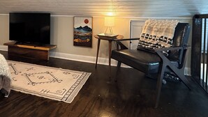A living room with a television on a wooden stand, a chair with a blanket, a small table with a lamp, and a rug with a geometric pattern. - Blissful Marsh Front GuestHouse Getaway! (Charleston)