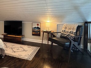 A living room with a television on a wooden stand, a chair with a blanket, a small table with a lamp, and a rug with a geometric pattern. - Blissful Marsh Front GuestHouse Getaway! (Charleston)