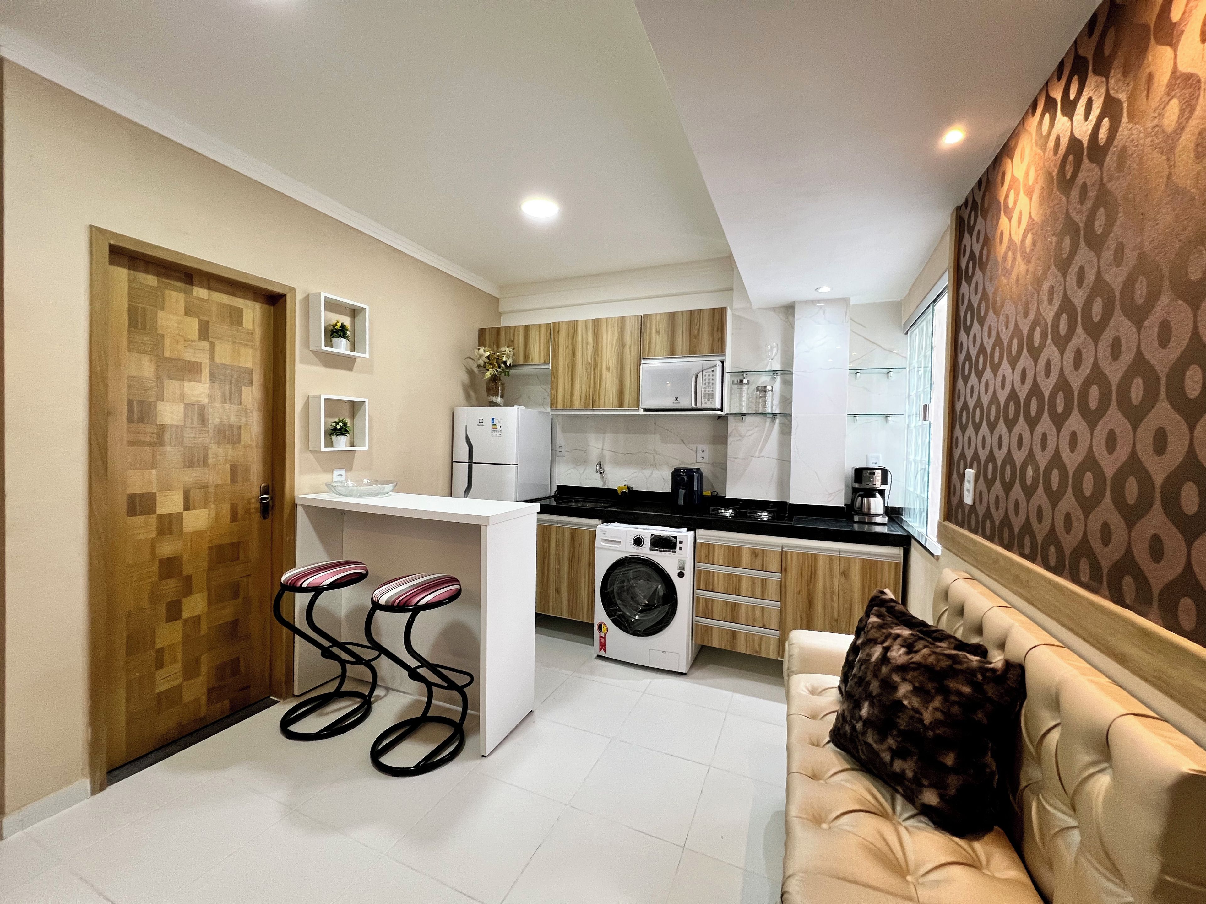A compact kitchen with a microwave, refrigerator, and washing machine. There is a bar stool and a sofa with a pillow.