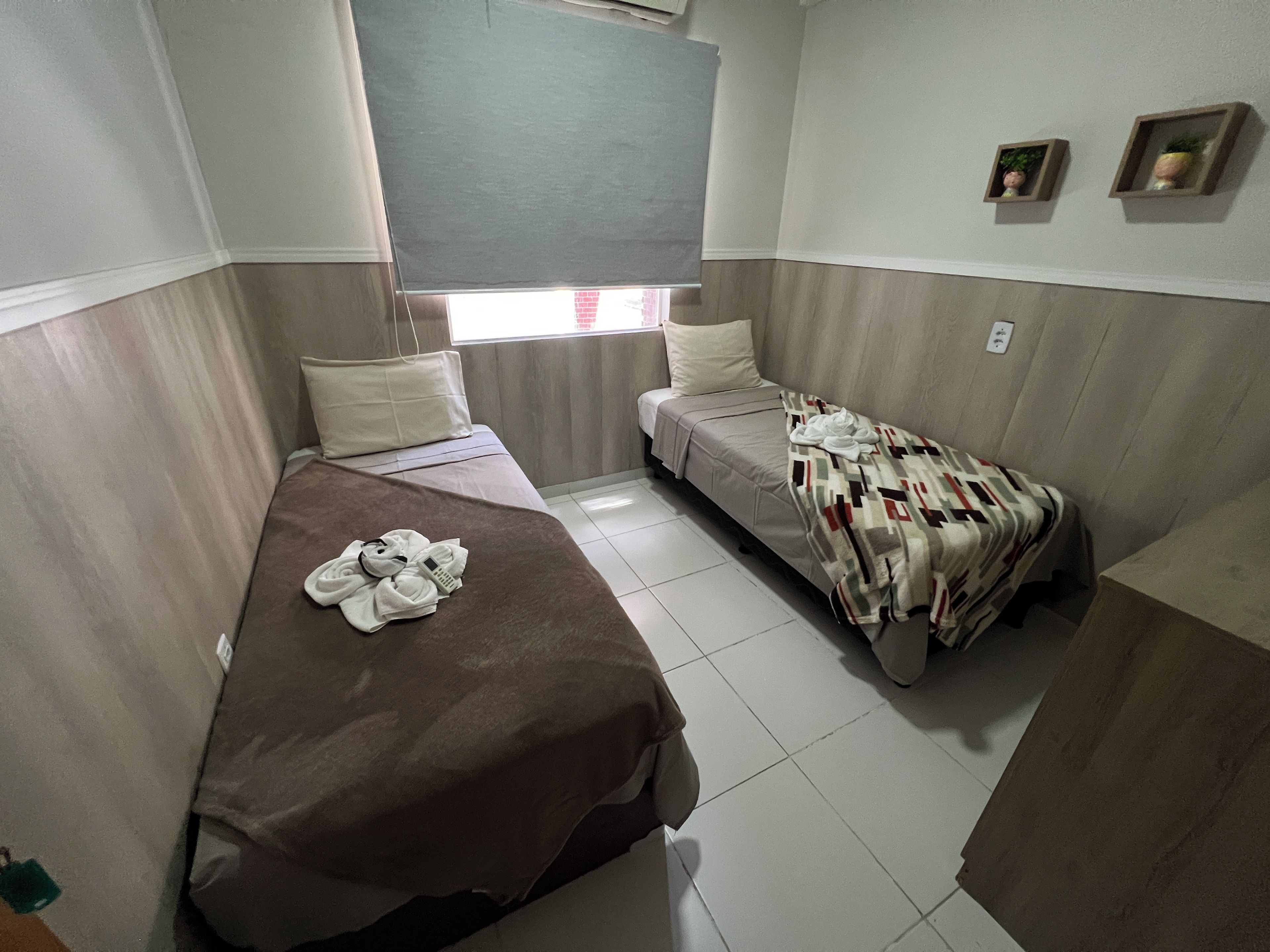 A small, compact room with two single beds, a window with a view, and a small shelf with decorative items.