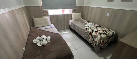 A small, compact room with two single beds, a window with a view, and a small shelf with decorative items.