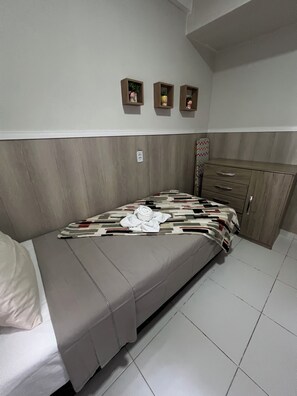 Una cama con una colcha estampada y una toalla enrollada, un cabecero de madera, un pequeño armario con cajones y un estante de pared con objetos decorativos.