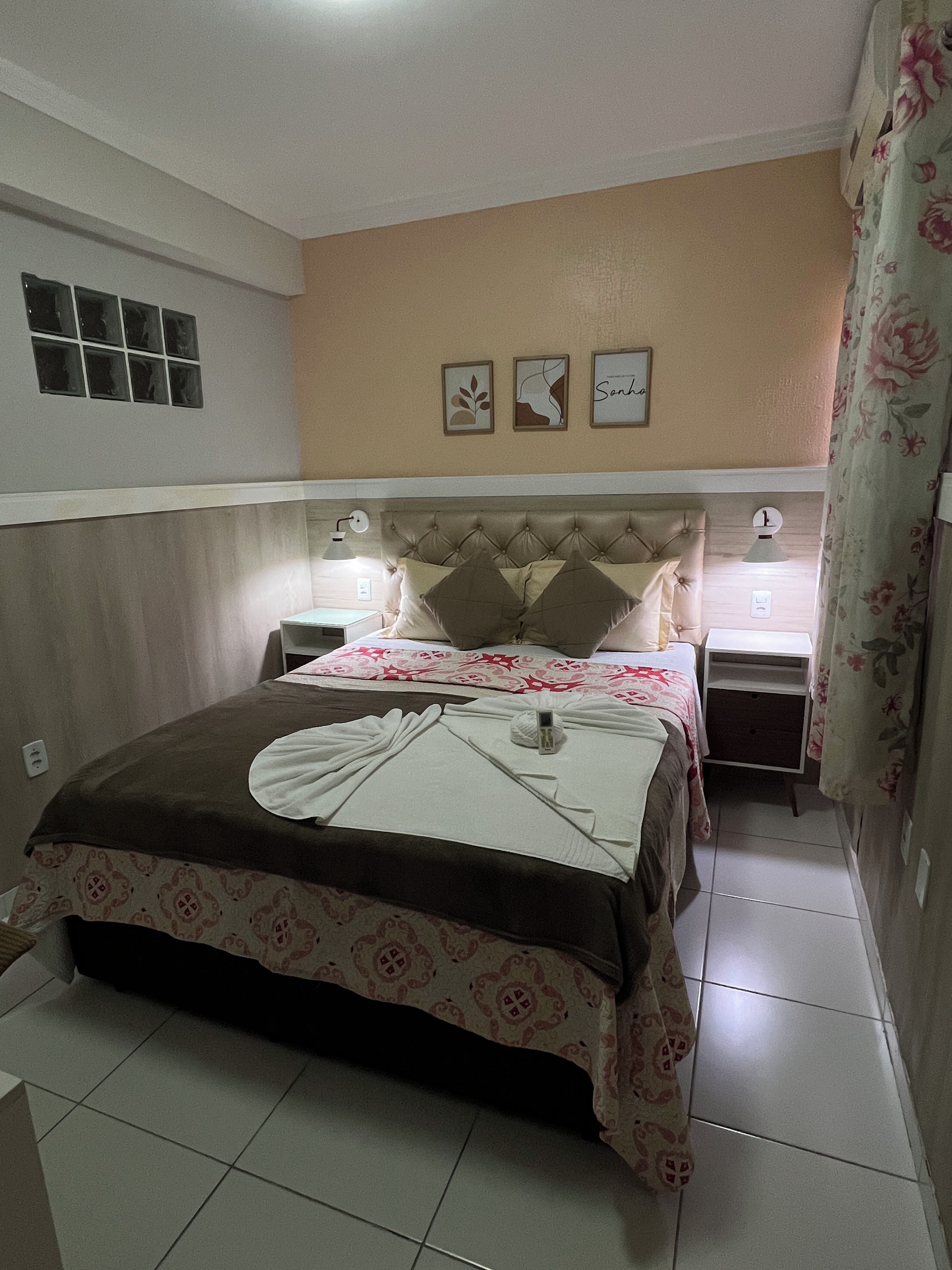 A bedroom with a bed, two bedside tables, a wall clock, and a window with blinds.