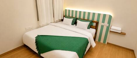 Una cama bien hecha con un plaid verde, una mesita de noche con lámpara y un estante en la pared con una toalla.