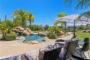 A pool area with lounge chairs and a table, surrounded by rocks and palm trees.