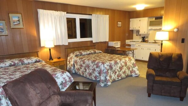 Una habitación con dos camas, un sillón marrón, una mesita, una kitchenette y una ventana con cortinas.