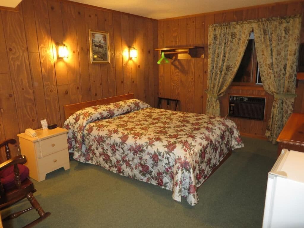 Een slaapkamer met een houten wand, een bed met bloemenbeddels, een nachtkastje, een schommelstoel en een radiator.