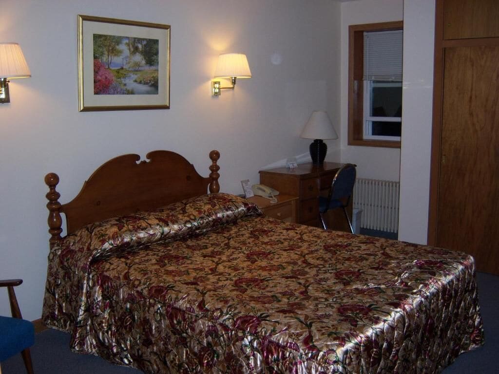 Ein Schlafzimmer mit einem hölzernen Bett, einem Nachttisch, einem Schreibtisch, einem Stuhl, einem Heizkörper und einem Fenster mit Jalousien.