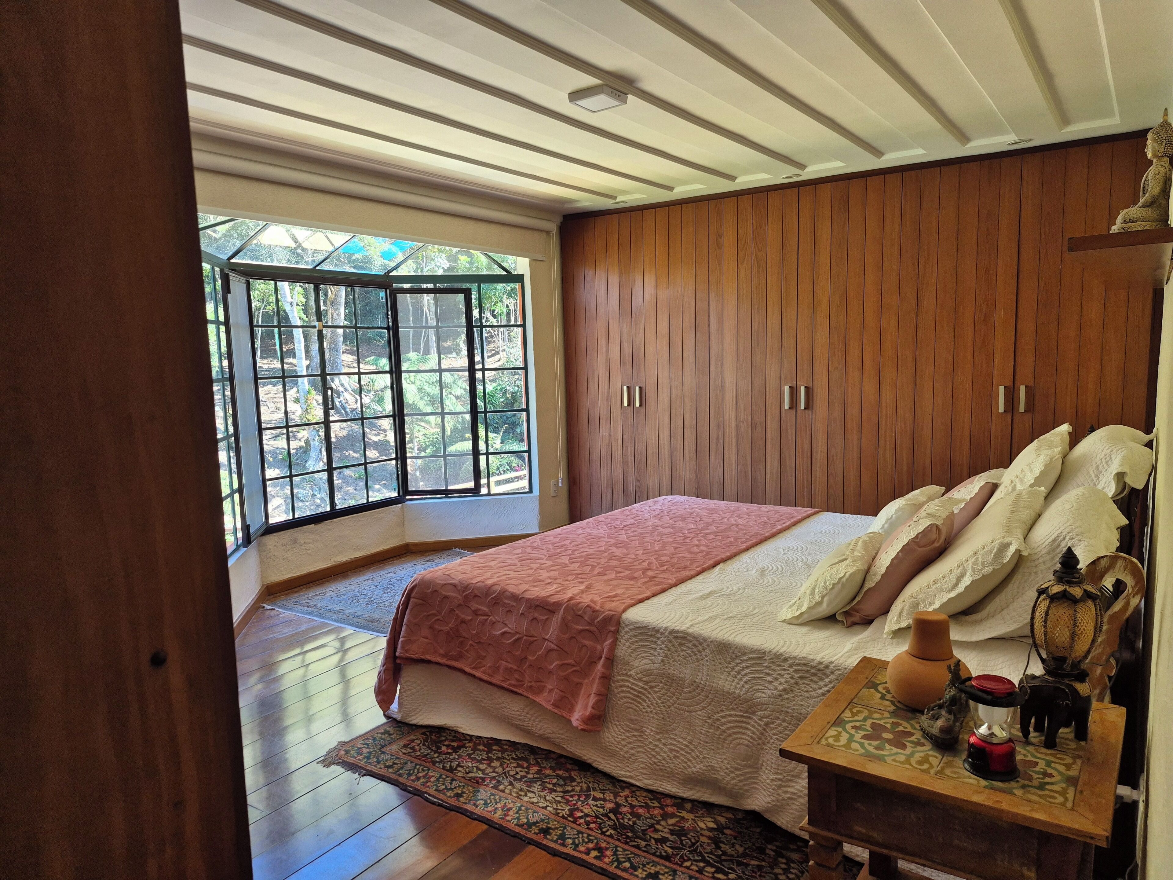 A bedroom with a large window, wooden wardrobe, and a bedside table with decorative items.