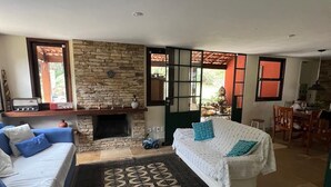 A spacious living area with a stone fireplace, a sofa, a patterned rug, and a dining area visible through a glass door. - English-style country house with stunning views. (Petrópolis)