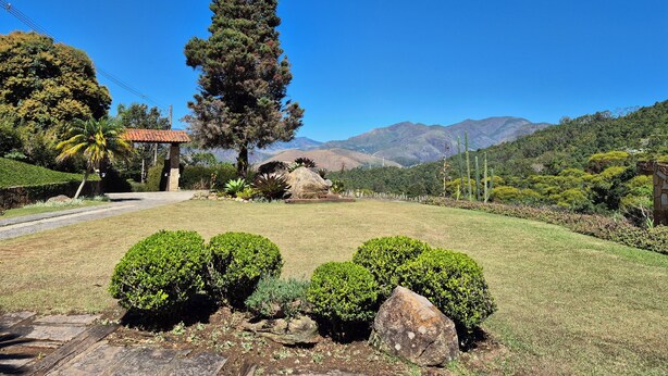A landscaped garden with a prominent tall tree, a small building, and a view of distant mountains. - English-style country house with stunning views. (Petrópolis)