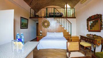 Quarto de hotel com uma cama, uma escrivaninha, uma cadeira, um espelho, uma decoração com o tema do sol, uma escada e um quadro na parede.