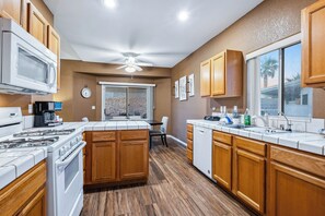 A modern kitchen with wooden cabinets, a central island, a dining area, and a view through a sliding glass door. - Cozy desert home near the Laughlin River (Bullhead City)