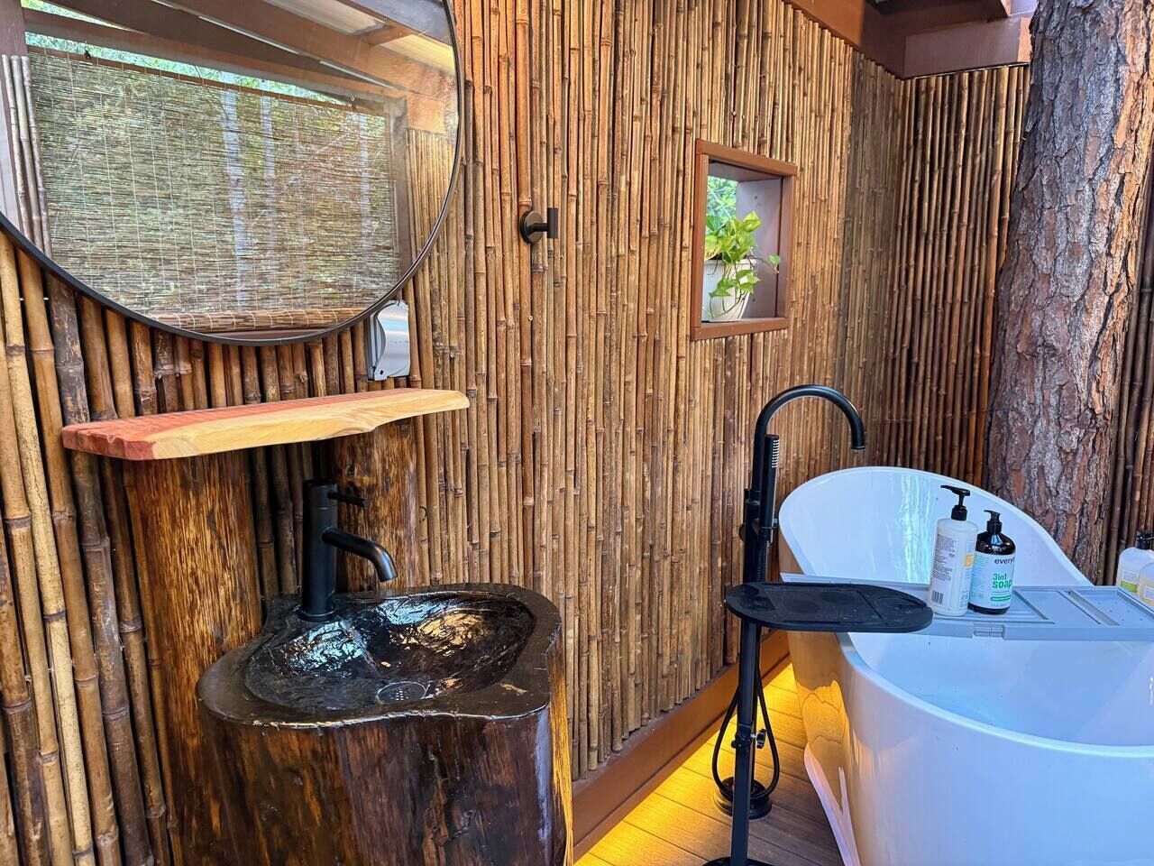 Ein Badezimmer mit Bambuswand, einem Holzregal, einem schwarzen Wasserhahn, einer weißen Badewanne und einem kleinen Fenster mit einer Pflanze.