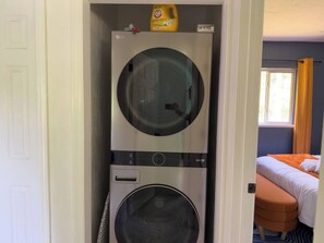 A laundry room with two stacked washing machines, a bottle of laundry detergent, and a bed with an orange blanket in the adjacent room. - Retro-Chic 4BR House w/ yard Downtown Gainesville (Gainesville)