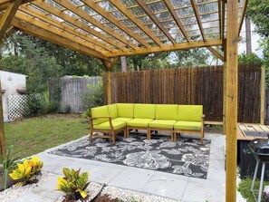 Ein überdachter Patio-Bereich mit einer Holzhütte, einem hellgrünen Ecksofa und einem gemusterten Teppich.