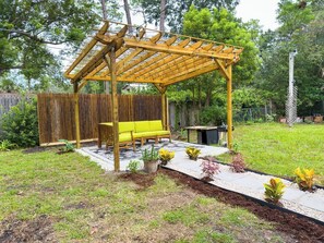 A wooden pergola with a yellow bench and a small table, surrounded by a garden with various plants and a wooden fence. - Retro-Chic 4BR House w/ yard Downtown Gainesville (Gainesville)