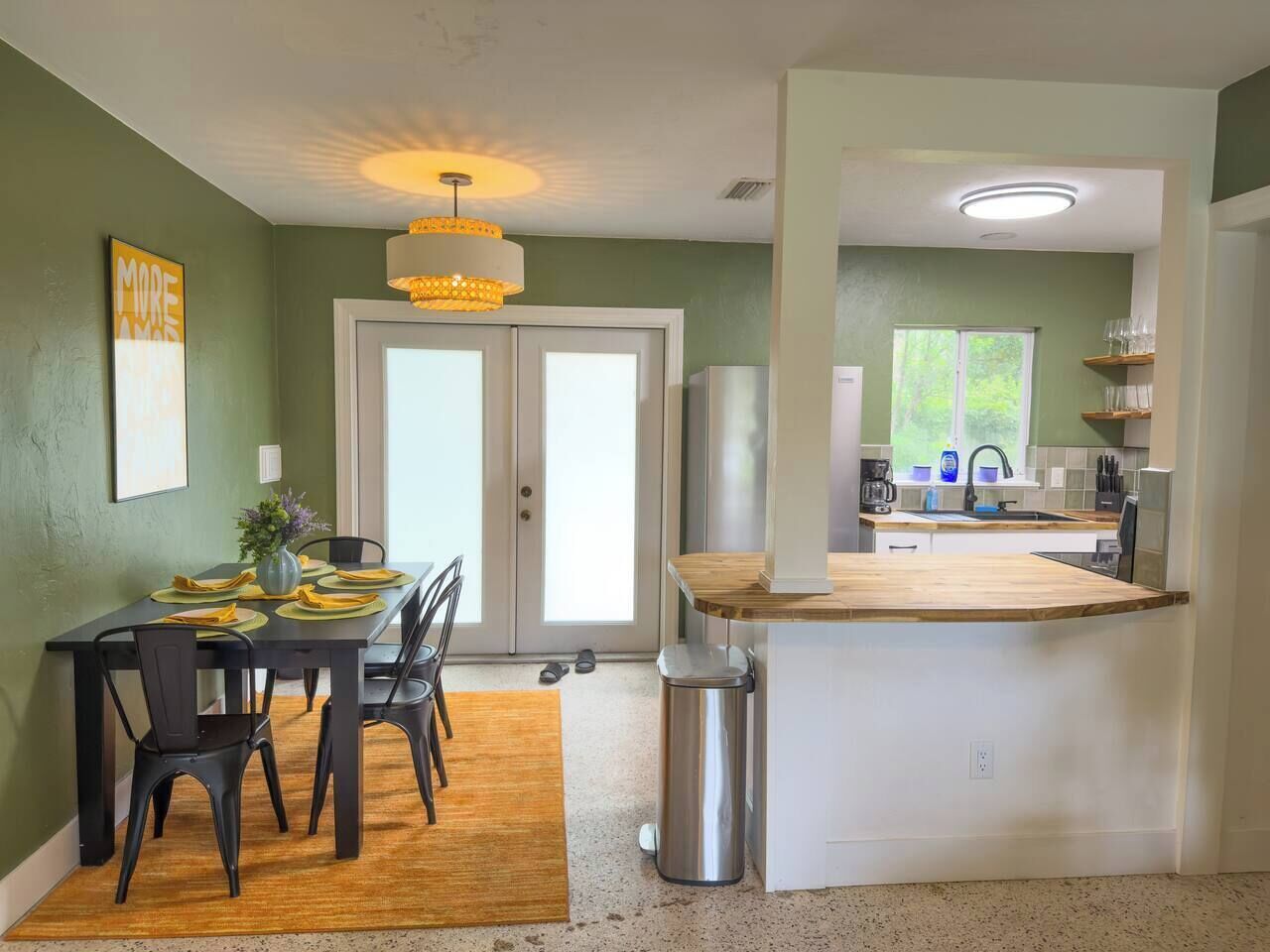 Un coin repas avec une table dressée pour quatre personnes, une cuisine dotée d’un plan de travail en bois et une poubelle.