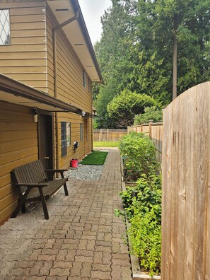 A wooden house with a paved patio, a bench, and a fenced garden area.