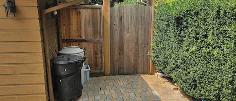 Un patio trasero con un cobertizo de madera, un bote de basura negro y un patio empedrado.