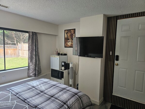 A bedroom with a bed, a TV, a heater, a microwave, and a window with a view of a backyard.