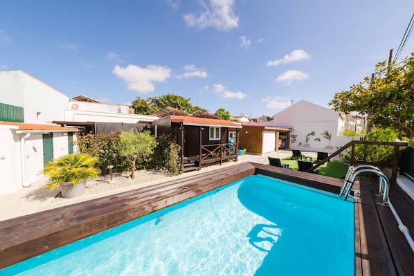 A swimming pool with a wooden deck, surrounded by buildings and greenery.