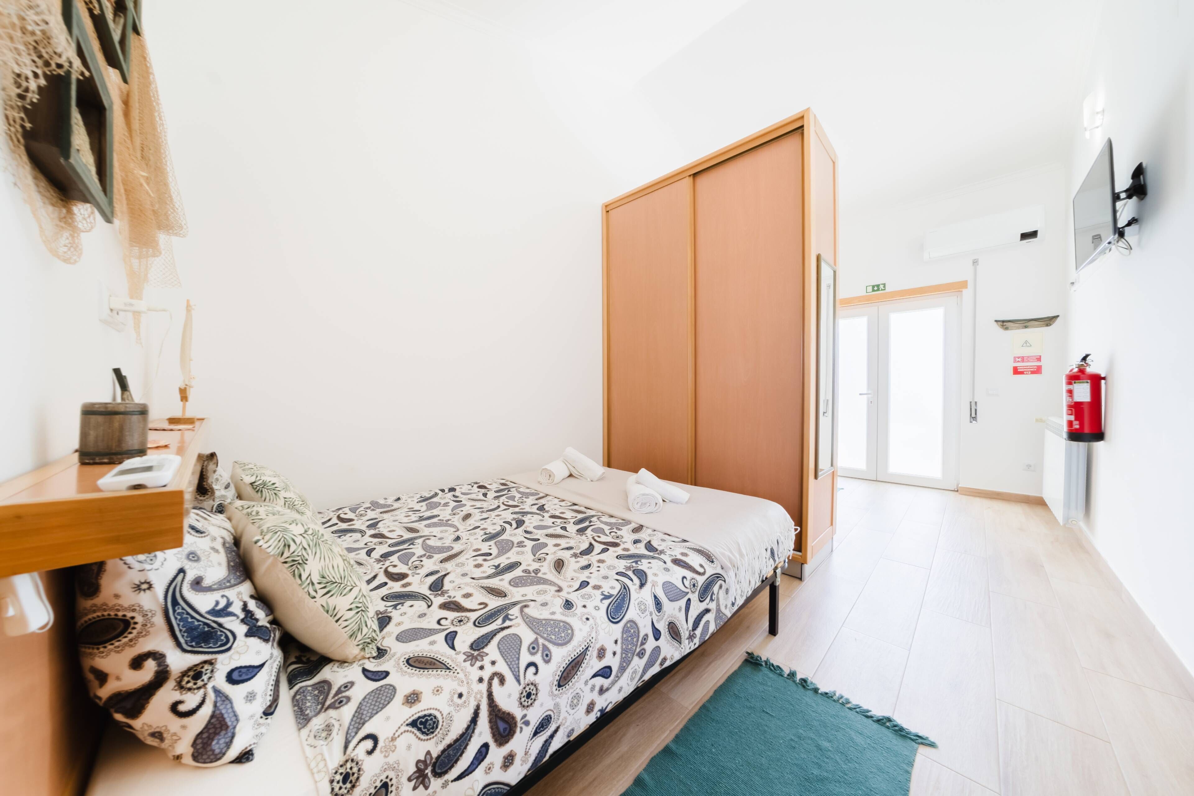 A hotel room with a bed, a desk, a chair, a small table, a fire extinguisher, and a door.