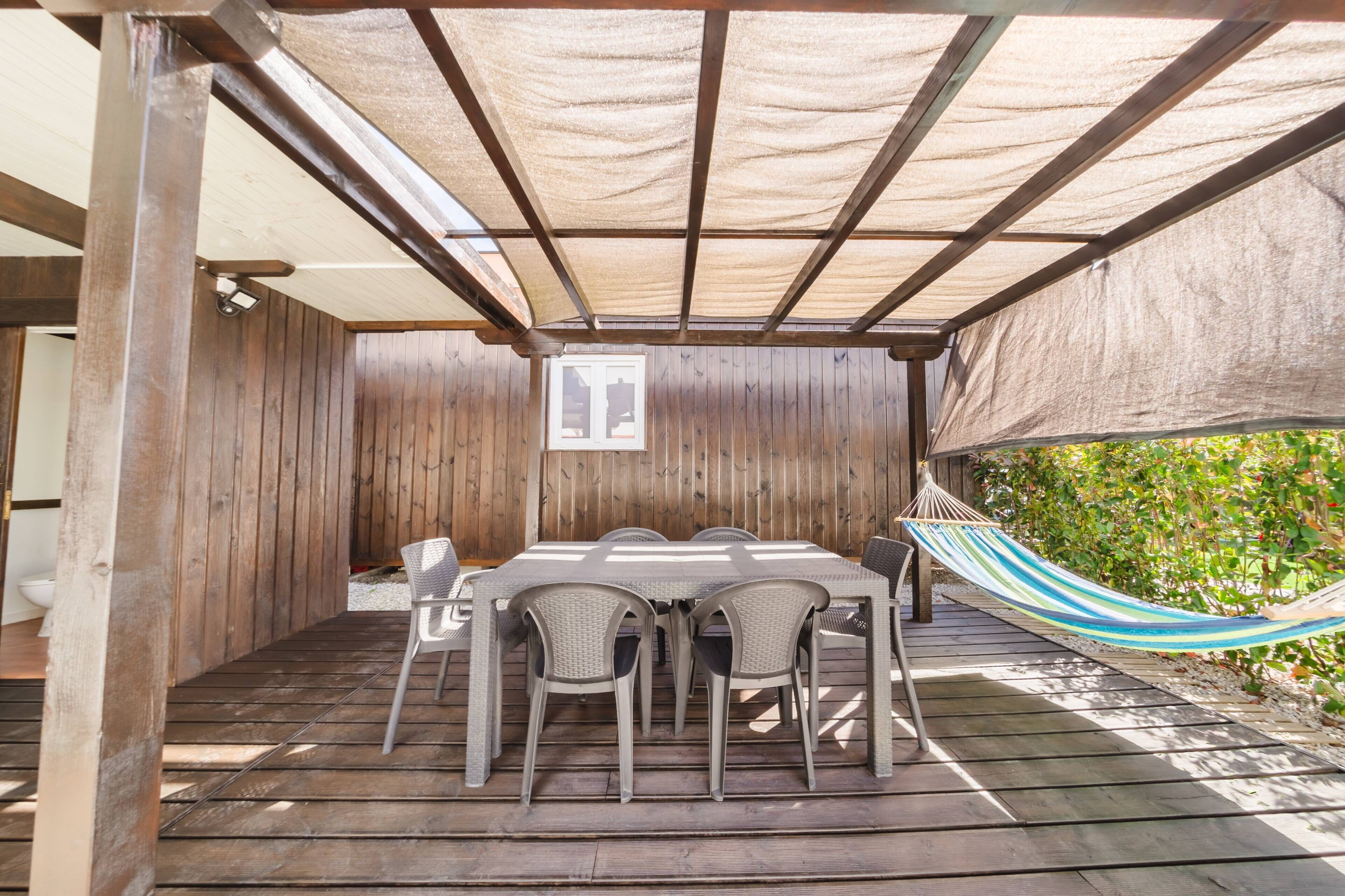 A wooden deck with a table and chairs, a hammock, and a pergola with a shade cloth.