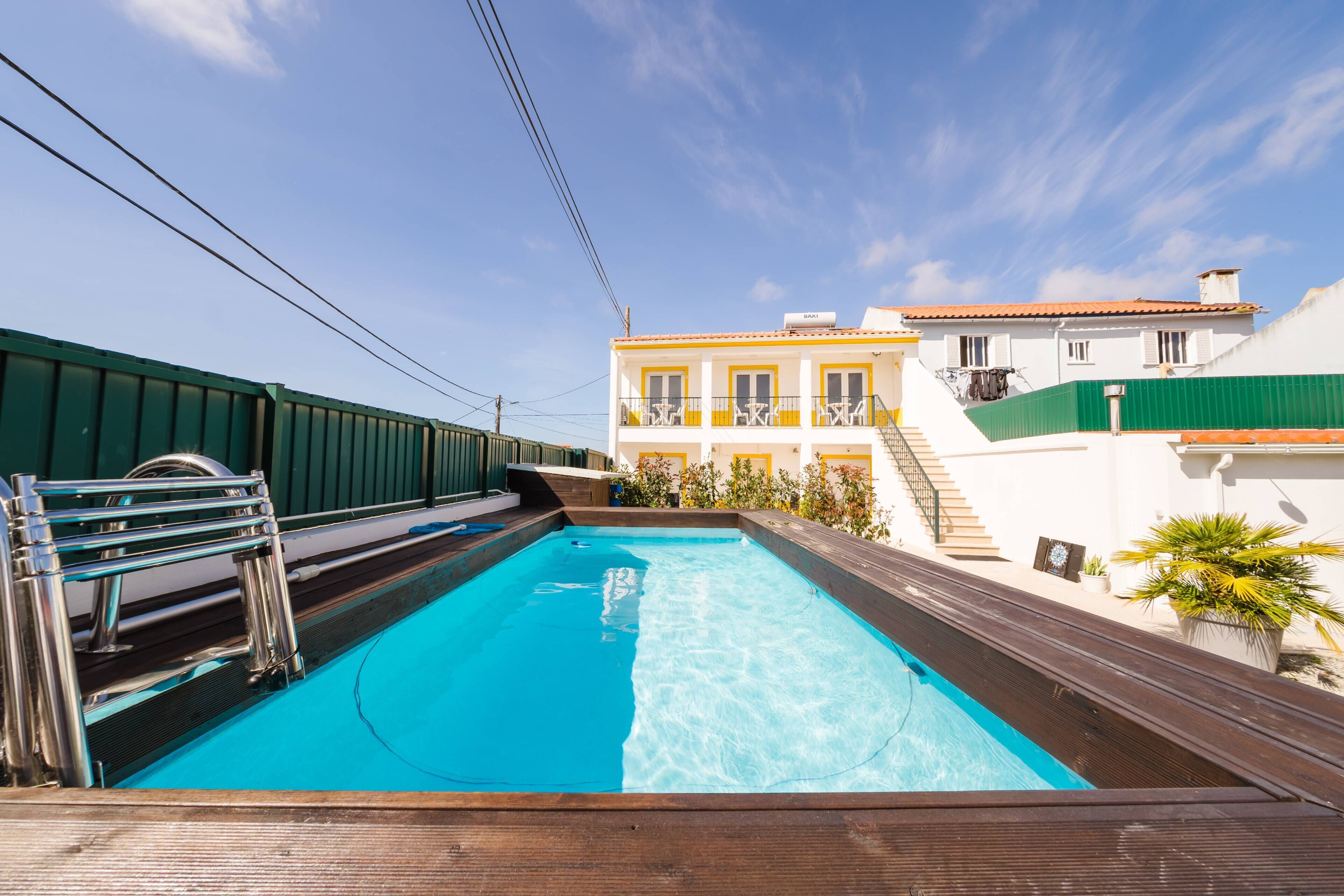 A swimming pool with a wooden deck, a metal ladder, and a building with balconies in the background.