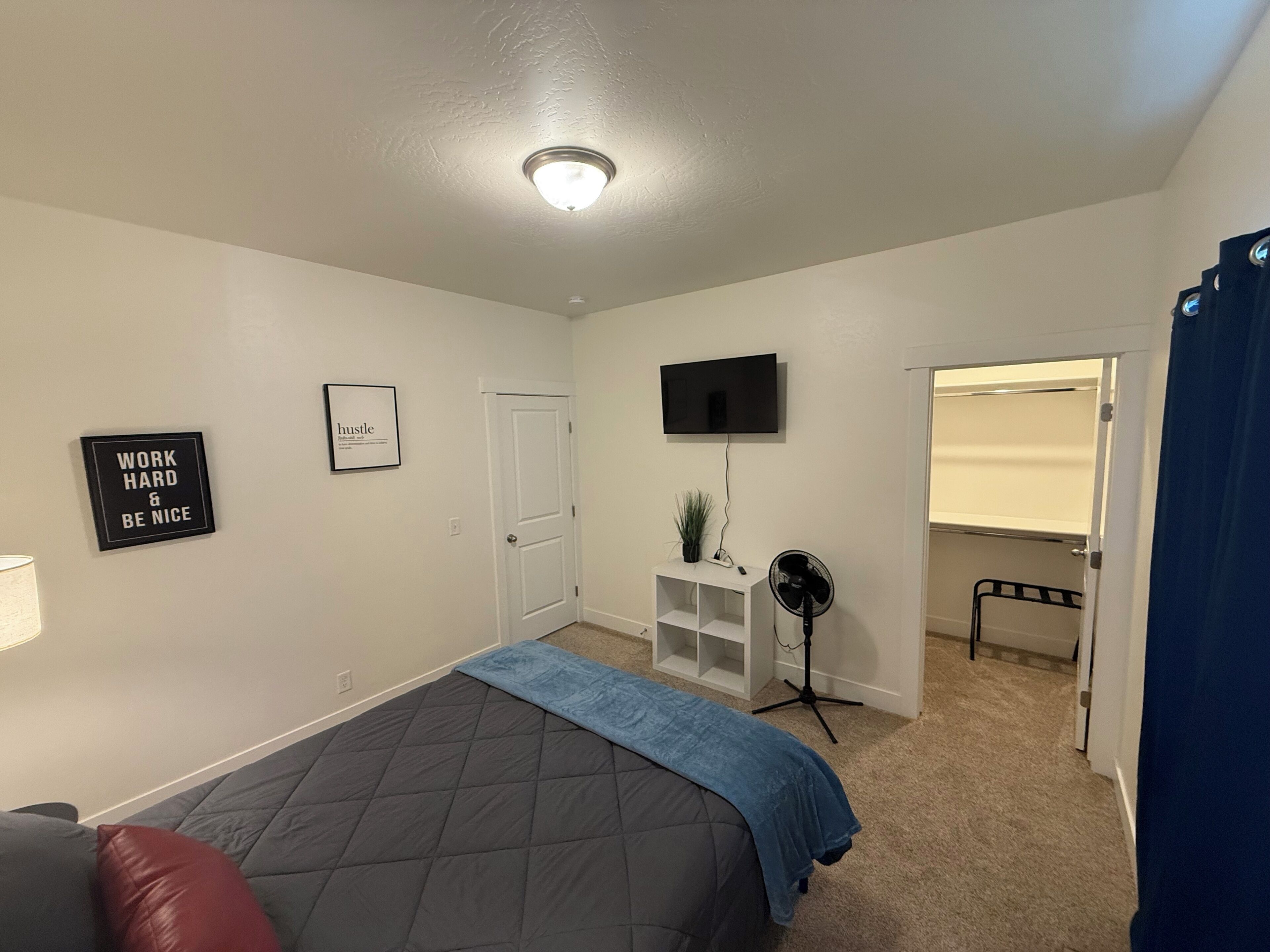 Un dormitorio con cama, televisor, ventilador, puerta y una pared decorada con carteles motivacionales.