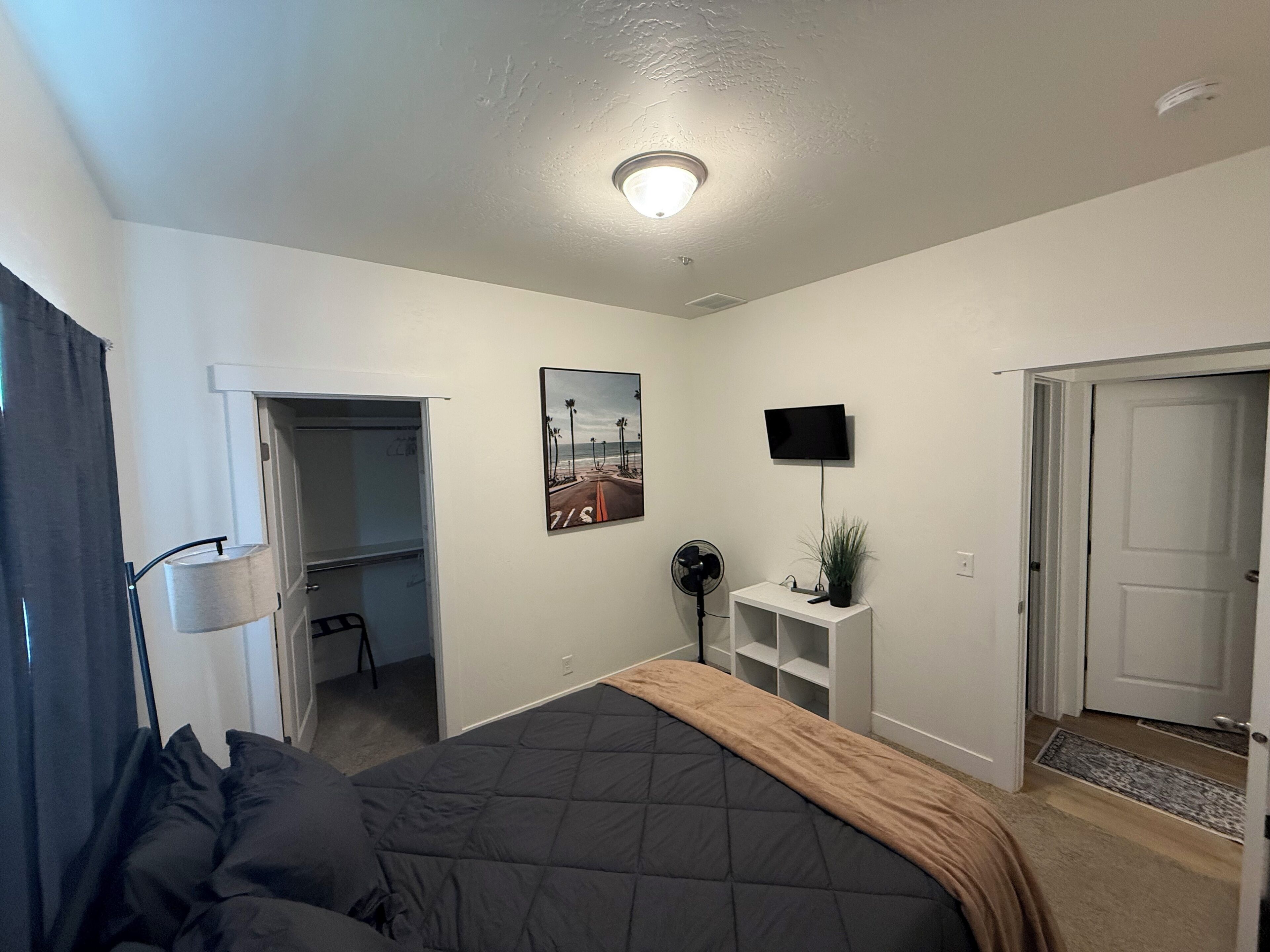 Une chambre à coucher avec un lit, un téléviseur fixé au mur, un ventilateur de plafond et une porte donnant sur une autre pièce.