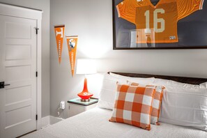 A bedroom with a bed, pillows, a framed jersey, a lamp, and two pennant flags. - 1BR Hot Tub Suite • Tennessee (Sevierville)