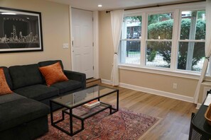 A living room with a sofa, a glass coffee table, a patterned rug, and a large window with a view of greenery. - Fully Furnished 2BR. 5-min walk to train. In-unit laundry. Parking Available (Chicago)