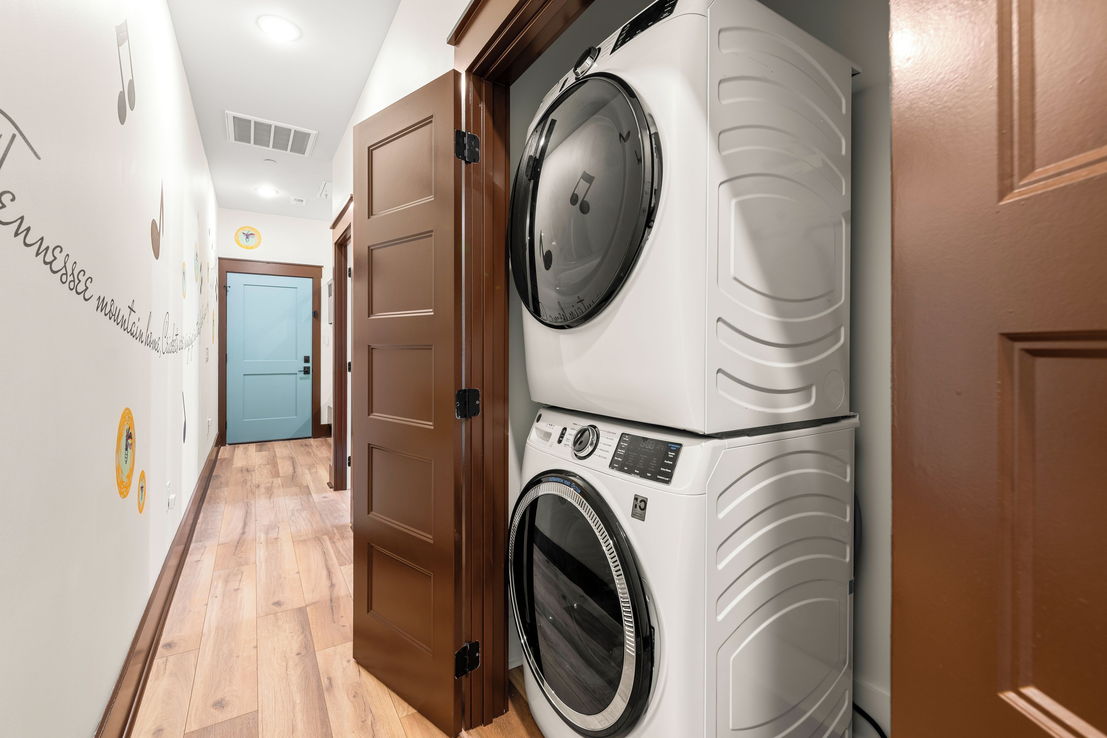 A laundry room with two stacked washing machines, wooden doors, and a hallway with a light blue door at the end.