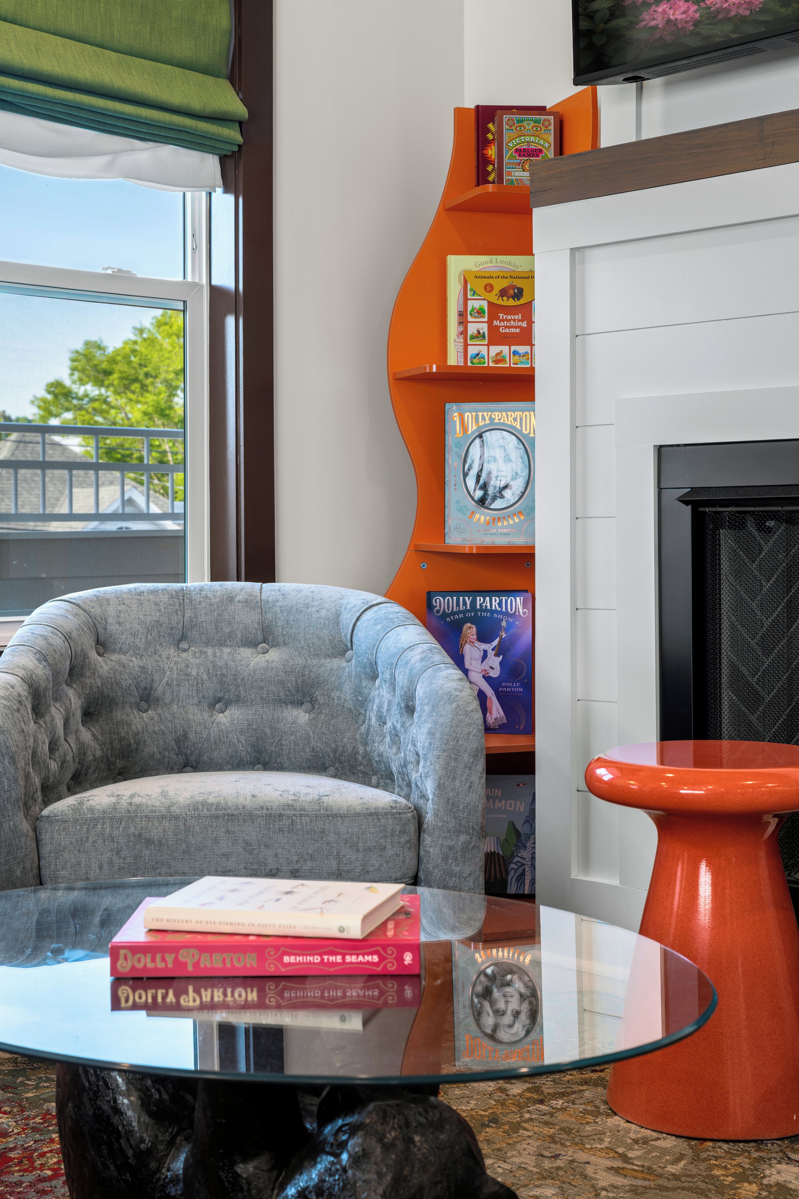 Una sala de estar con un sillón gris, una mesa de centro de vidrio con libros, una estantería naranja y una chimenea.