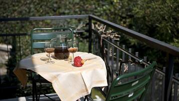 Un tavolo con una tovaglia, due bicchieri da vino, una bottiglia e un oggetto rosso su un balcone con sedie verdi e una ringhiera.