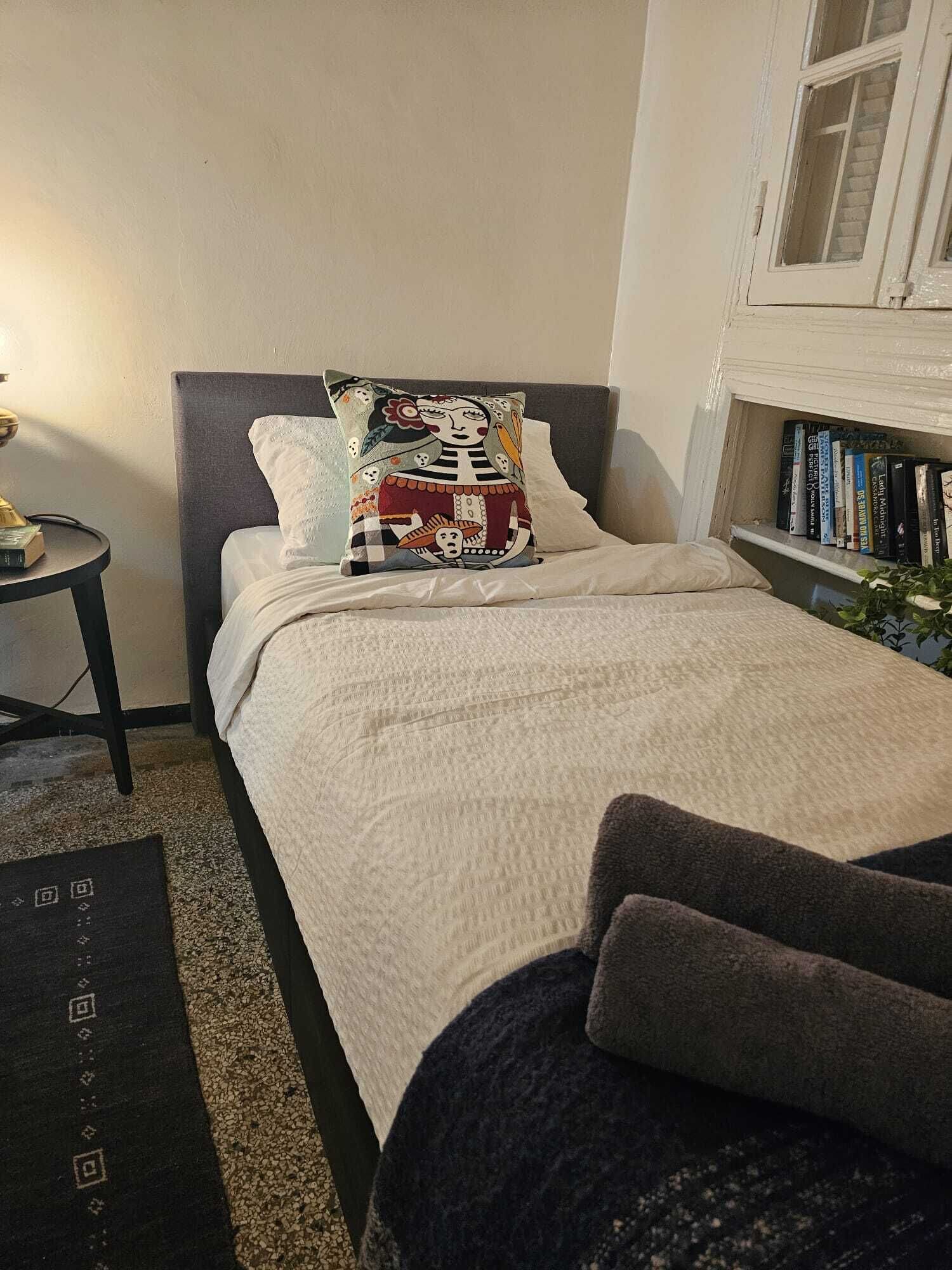Une chambre à coucher avec un lit, des oreillers, une table de chevet, une lampe, une bibliothèque et une fenêtre.