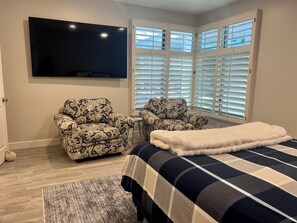 A bedroom with a bed, two patterned armchairs, a small side table, a television mounted on the wall, and a window with blinds. - Welcome to your dream retreat in Lantern Bay Villas (Dana Point)