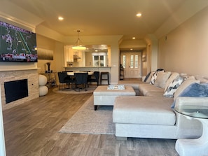 A modern living room with a fireplace, a TV mounted on the wall, a dining area with a kitchen in the background, and a large sofa.