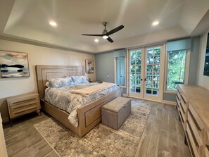 A bedroom with a bed, a nightstand, a dresser, a ceiling fan, and a large window with a view of greenery. - Welcome to your dream retreat in Lantern Bay Villas (Dana Point)
