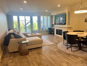 Ein modernes Wohnzimmer mit Sofa, Hocker, Couchtisch und einem Essbereich mit Tisch und Stühlen. Ein Kamin und ein an der Wand befestigter Fernseher sind ebenfalls vorhanden.