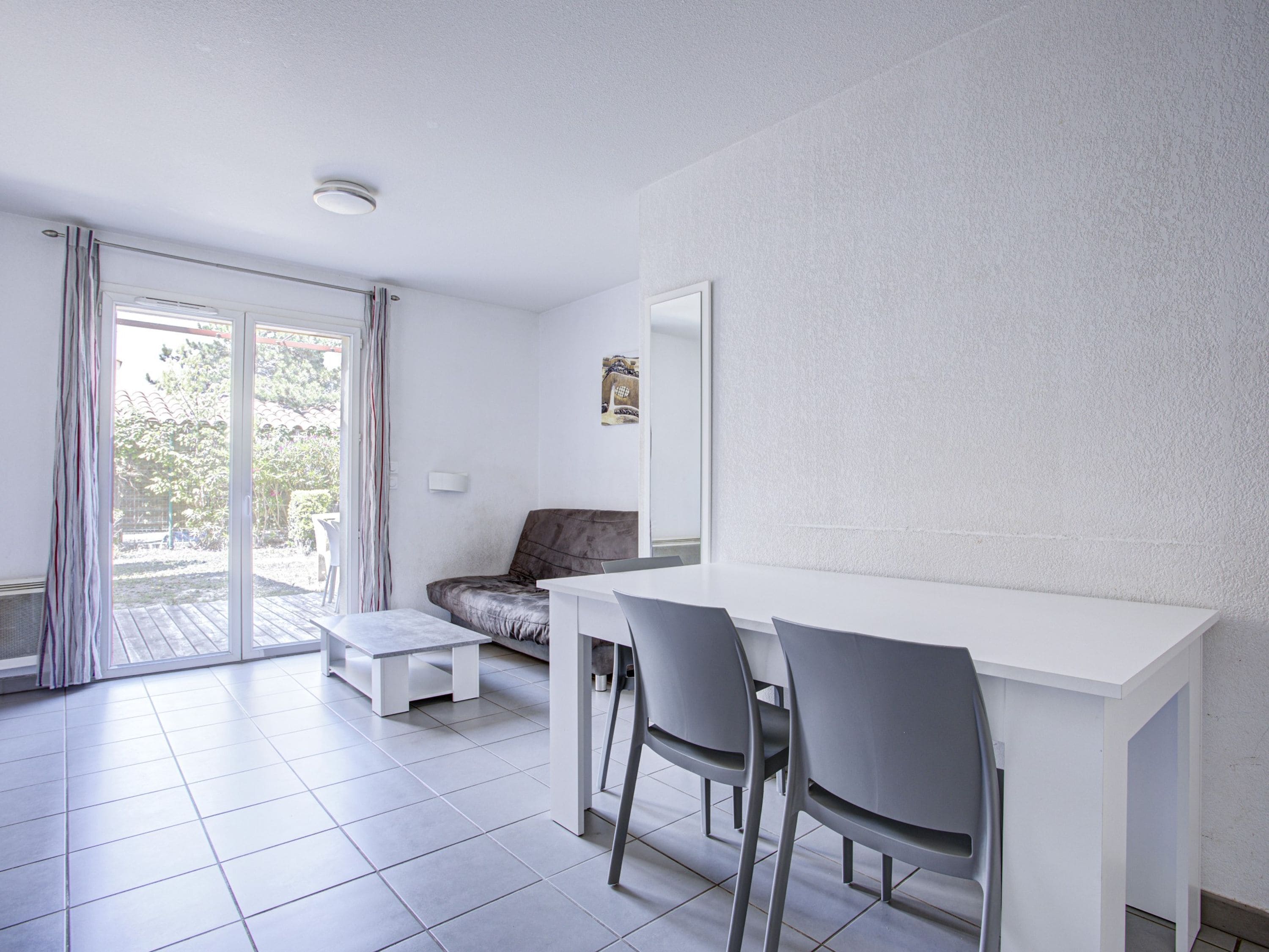 Une chambre moderne avec un bureau blanc, deux chaises, un canapé, une petite table et une porte-fenêtre coulissante donnant sur un espace extérieur.