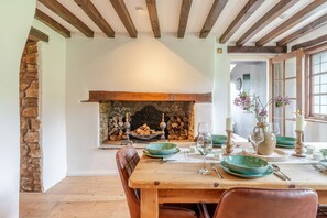 A dining area with a wooden table set for four, a stone fireplace, and a view of the outside through a window. - 1641 Historic Farmhouse Sleeps 16 Bath Bristol (Bristol)