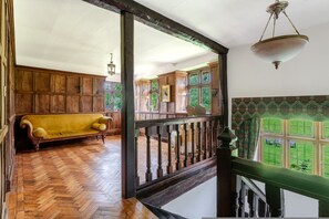 A spacious room with a herringbone wooden floor, a yellow sofa, and a view of a garden through large windows. - 1641 Historic Farmhouse Sleeps 16 Bath Bristol (Bristol)