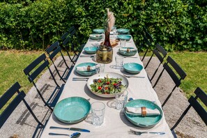 Une longue table à manger installée à l’extérieur, avec des chaises, des assiettes, des verres et un centre de table.