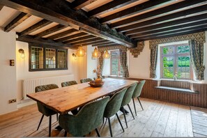 A dining area with a wooden table, green upholstered chairs, exposed wooden beams, and a window with patterned curtains. - 1641 Historic Farmhouse Sleeps 16 Bath Bristol (Bristol)