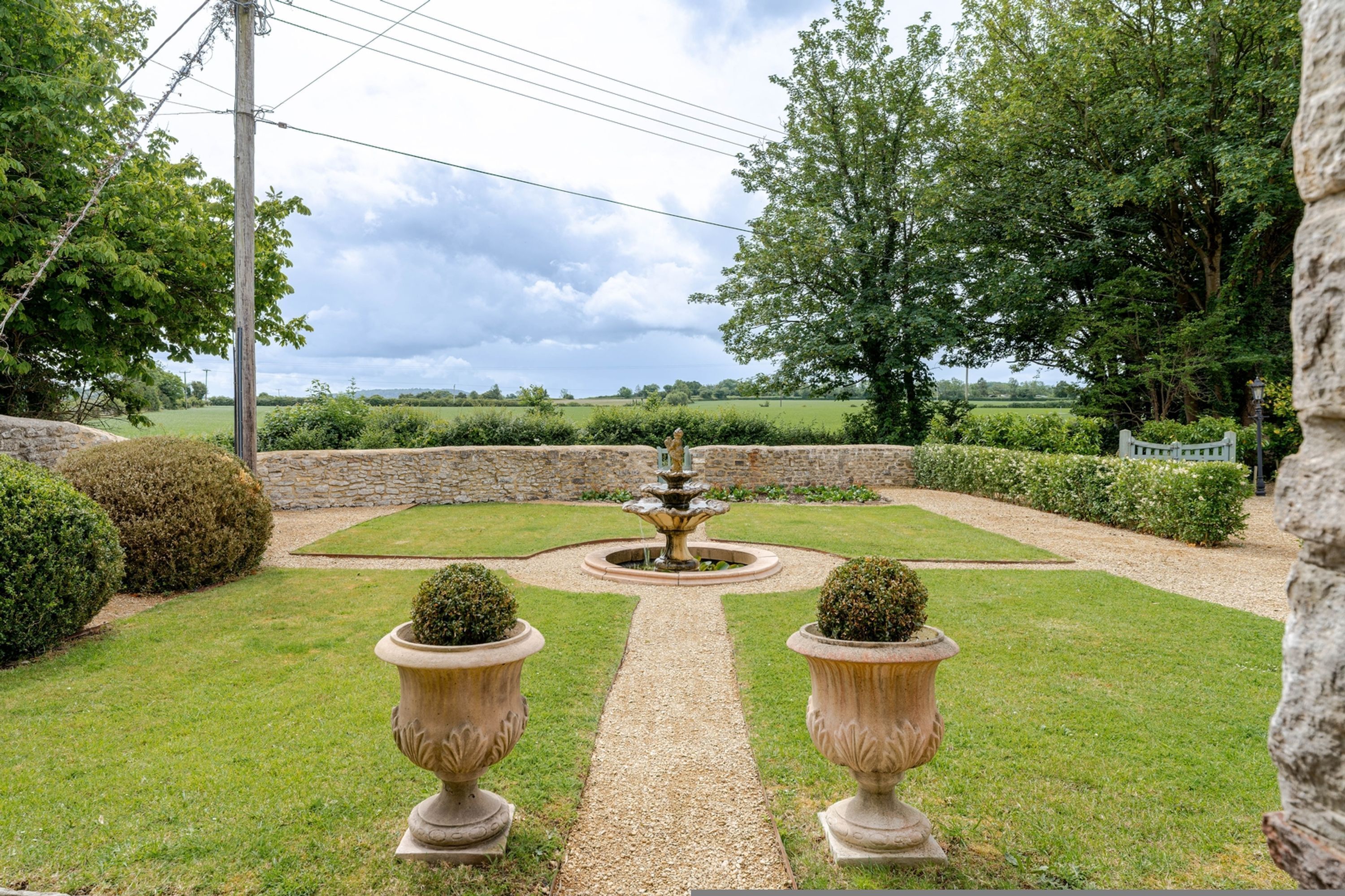 A garden with a central fountain, surrounded by neatly trimmed hedges and a stone wall.